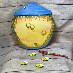 Può includere: Un barattolo di miele di legno con un coperchio blu e miele giallo che cola lungo i lati. Ci sono cinque piccole api di legno sparse intorno al barattolo.