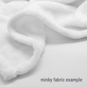 May include: Close-up of a white minky fabric. The fabric is soft and plush, with a fuzzy texture. Minky fabric is often used for blankets, throws, and other soft goods.