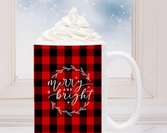 May include: White ceramic mug with a red and black buffalo plaid pattern. The mug features the words "merry and bright" in a white script font, encircled by a wreath. The mug is filled with whipped cream.