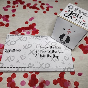 May include: A white box with the text "Give you a hug" on the front. The box has a flap that can be pulled to reveal a message inside. The box is surrounded by red and pink confetti.