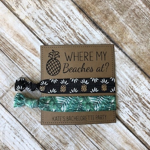 May include: Two hair ties with tropical prints. One is black with gold pineapples and white flowers. The other is green with palm leaves. The hair ties are on a brown card with the text "Where my Beaches at?" and "Kate's Bachelorette Party".