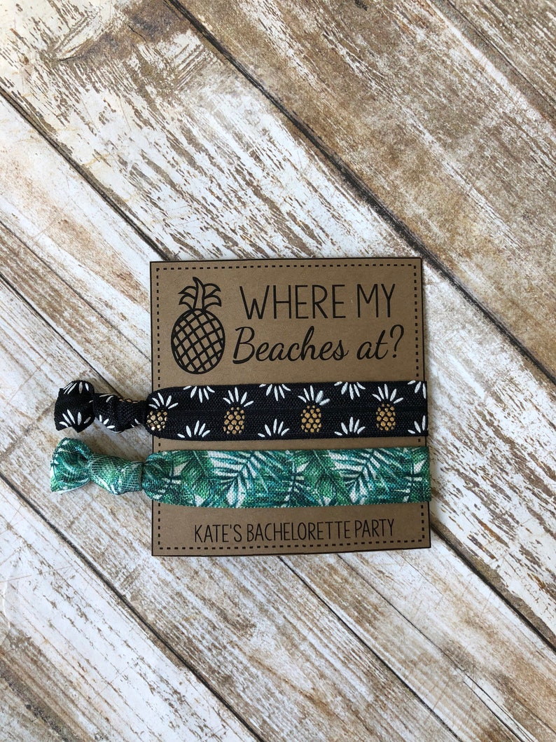 May include: Two hair ties with tropical prints. One is black with gold pineapples and white flowers. The other is green with palm leaves. The hair ties are on a brown card with the text "Where my Beaches at?" and "Kate's Bachelorette Party".