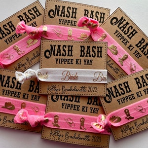 May include: Pink and white hair ties with gold foil accents. The hair ties have a western theme with boots and hats. The hair ties are packaged with brown cards that say "Nash Bash Yippie Ki Yay" and "Kelly's Bachelorette 2023".