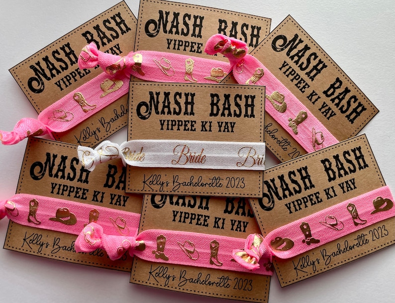 May include: Pink and white hair ties with gold foil accents. The hair ties have a western theme with boots and hats. The hair ties are packaged with brown cards that say "Nash Bash Yippie Ki Yay" and "Kelly's Bachelorette 2023".