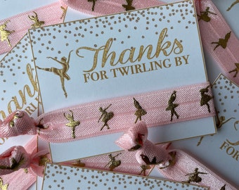 May include: Pink elastic hair ties with gold ballerina silhouettes printed on them. Each hair tie has a white card attached with gold text that reads "Thanks for twirling by".