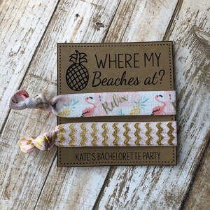 May include: Two pink and gold hair ties with a brown card that says "Where my Beaches at?" and "Kate's Bachelorette Party". The hair ties have a flamingo and pineapple design.