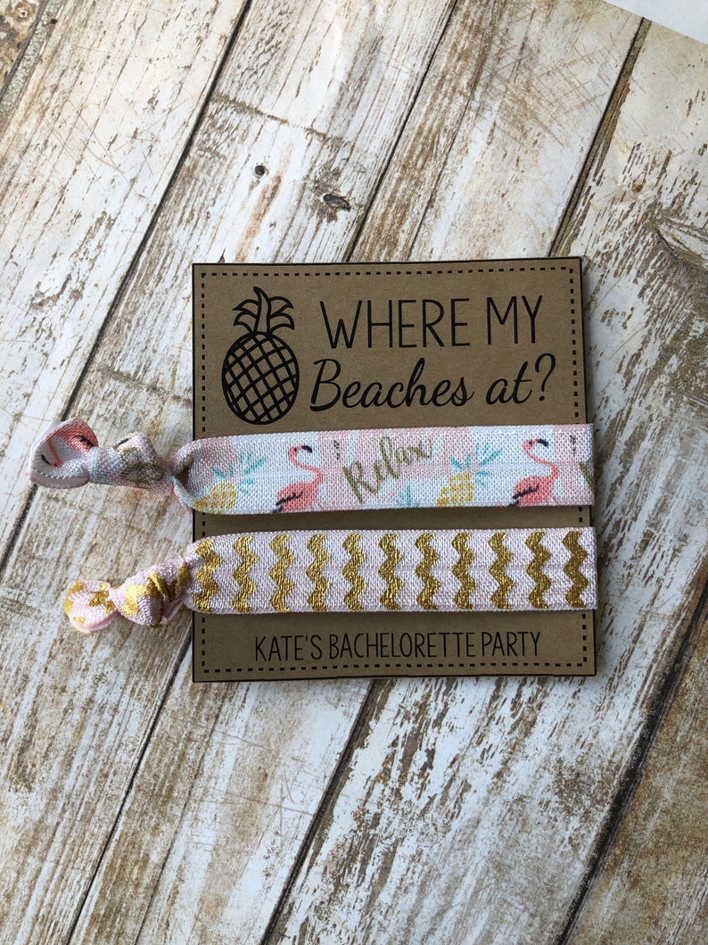 May include: Two pink and gold hair ties with a brown card that says "Where my Beaches at?" and "Kate's Bachelorette Party". The hair ties have a flamingo and pineapple design.
