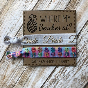 May include: Two colorful bracelets with pineapple designs and the text "Where my beaches at?" on a brown card. The top bracelet is white with gold text that says "Bride". The bottom bracelet is multi-colored with a pineapple pattern. The card also says "Kate's Bachelorette Party".