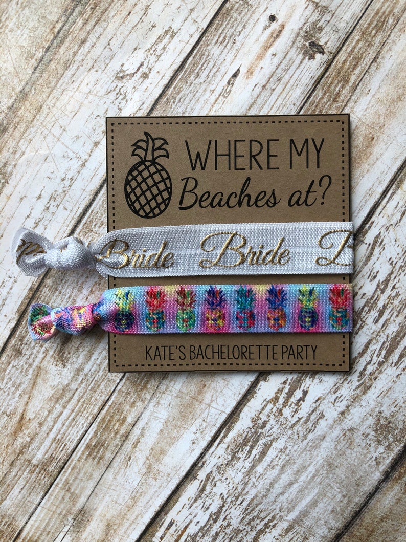 May include: Two colorful bracelets with pineapple designs and the text "Where my beaches at?" on a brown card. The top bracelet is white with gold text that says "Bride". The bottom bracelet is multi-colored with a pineapple pattern. The card also says "Kate's Bachelorette Party".