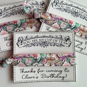 May include: A set of tea-themed party favors with a white background and black text that reads "You are my cup of tea". The favors include a colorful fabric hair tie with a teapot and teacup design and a thank you tag that reads "Thanks for coming to Clare's Birthday!"