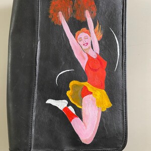 May include: Black leather clutch bag featuring a painted image of a cheerleader in a red and yellow uniform, holding pom-poms and jumping in the air.
