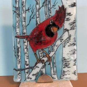 May include: A rectangular fused glass art piece featuring a vibrant red cardinal perched on a white birch tree branch. The background depicts a light blue sky and additional white birch trees with black markings, all supported by a stone base.