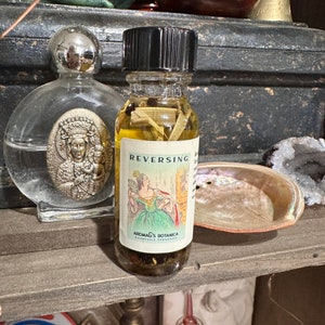 May include: A small glass bottle with a black cap filled with a yellow liquid and herbs. The label reads "REVERSING" and "Aromas' Botanica".