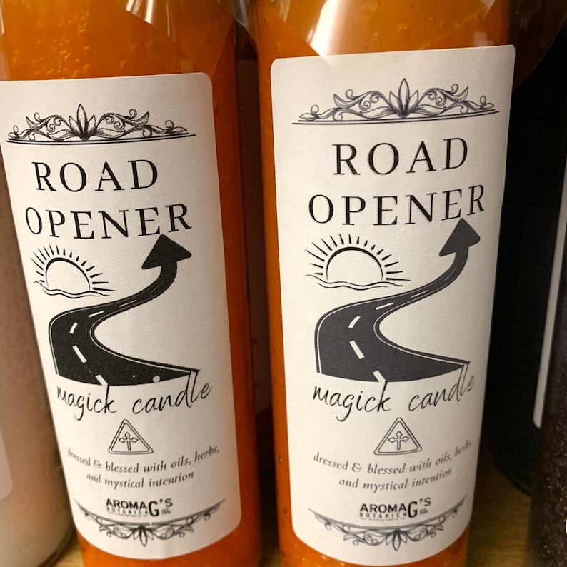 May include: Two orange glass candle jars with white labels. The labels have black text that reads "Road Opener magick candle" and a black and white illustration of a road with a sun and an arrow. The labels also have a small black and white illustration of a cross with a circle around it. The text below the illustration reads "dressed & blessed with oils, herbs, and mystical intention." The text at the bottom of the label reads "AromaG's Botanica."