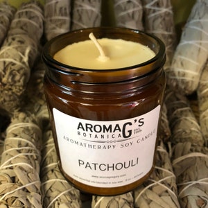 May include: A brown glass jar candle with a white label that reads "Aroma G's Botanica Est. 1999 Aromatherapy Soy Candle Patchouli www.aromagrigory.com 6 oz - 40 hours Soy essential oils blended in soy wax".