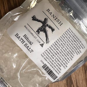 May include: A clear plastic bag containing ritual bath salt. The label reads "BANISH" with a graphic of a figure. Text on the label includes "Ritual BATH SALT" and "MAGICKAMULET.COM". The label also includes instructions for use.