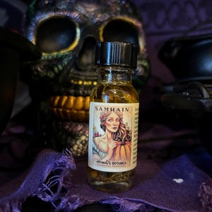 May include: A small glass bottle of Samhain oil with a black cap, featuring a label with a woman and pumpkin illustration. The bottle is in front of a decorative skull and purple fabric. The text "Aromag's Botanica" is visible.