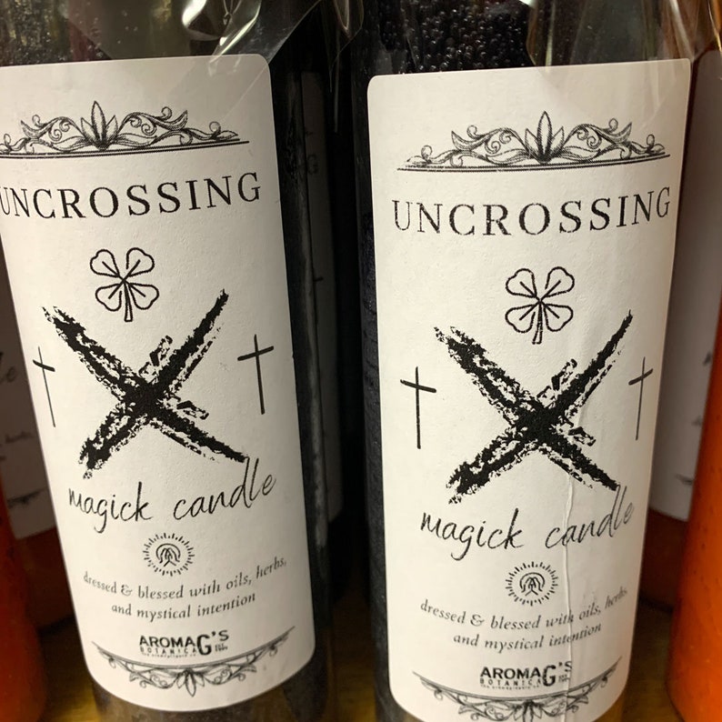 K&ouml;nnte beinhalten: Zwei schwarze Glaskerzen mit wei&szlig;en Etiketten. Die Etiketten zeigen ein vierbl&auml;ttriges Kleeblatt, durchgestrichene Kreuze und den Text "UNCROSSING magick candle". Der Text unter dem Kerzennamen lautet "dressed & blessed with oils, herbs, and mystical intention". Am unteren Rand des Etiketts steht der Text "AROMAG'S BOTANICALS".