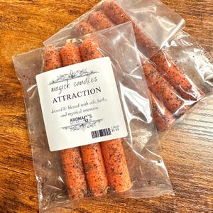 May include: Three orange candles with a brown speckled finish are packaged in a clear plastic bag. The label reads "magick candles ATTRACTION dressed & blessed with oils, herbs, and mystical intention AROMAG'S BOTANICALS 13639 $5.99"
