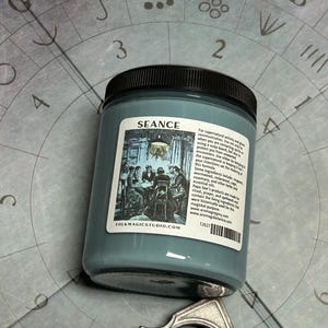 May include: A teal-colored jar with a black lid, labeled "SEANCE." The label features an illustration of people gathered around a table. The jar is set against a gray background with letters and numbers.