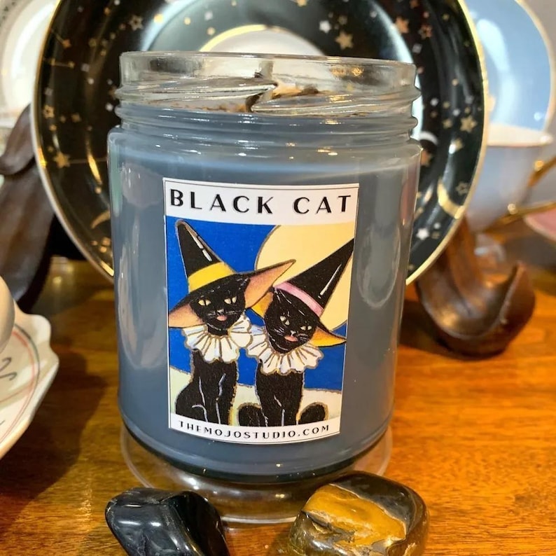 Może przedstawiać: Szklany słoik na świecę w kolorze szarym z ilustracją czarnego kota na etykiecie. Etykieta zawiera napis "Black Cat" i "The Mojo Studio.com".