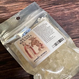 May include: A clear plastic bag containing bath salts. The label reads "DEVIL BEGONE" with an illustration of two figures. The text "AROMAG'S BOTANICA NASHVILLE TENNESSEE" is also visible. The bag is partially transparent, showing the crystalline contents.