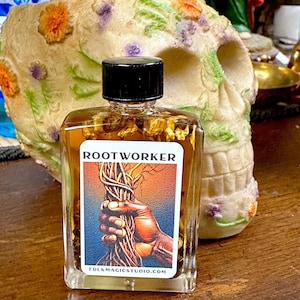 May include: A clear, square glass bottle with a black cap, labeled "ROOTWORKER." The label features a hand holding roots. The bottle is filled with a golden liquid and small pieces of plant matter. A decorative skull is in the background.
