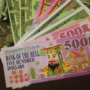 May include: A collection of various paper currencies, including U.S. dollars and bills from the Bank of the Hell. The bills feature colorful designs and text, with denominations visible. Some bills are fanned out, showcasing the variety of notes.