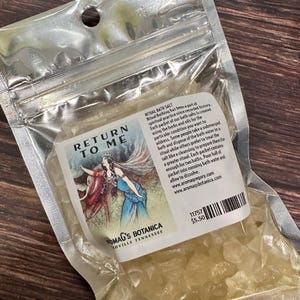 May include: A clear plastic bag filled with bath salts. The label reads "RETURN TO ME" with an illustration of two figures. Text on the label describes the product as ritual bath salts, with instructions for use. The brand name is NOMAG'S BOTANICA.