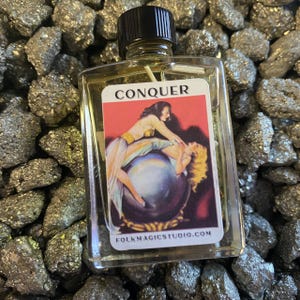 May include: A small glass bottle with a black cap containing a yellow liquid. The label on the bottle reads "CONQUER" and features a vintage illustration of two women on a globe. The website "folkmagicstudio.com" is printed at the bottom of the label.