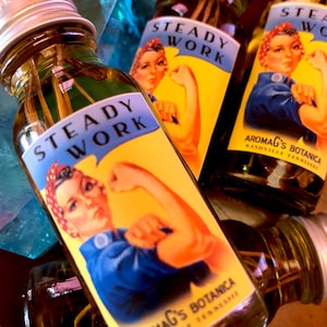 May include: Three glass bottles with yellow labels and a vintage-style illustration of a woman with her arm raised. The label reads "STEADY WORK" and "AromAg's Botanica Nashville, Tennessee".