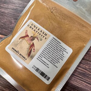 May include: A clear plastic bag filled with powdered incense. The label reads "UNLEASH CREATIVITY" and features a vintage illustration. The brand name "AROMAG'S BOTANICA" is also visible. The bag contains a light brown powder.