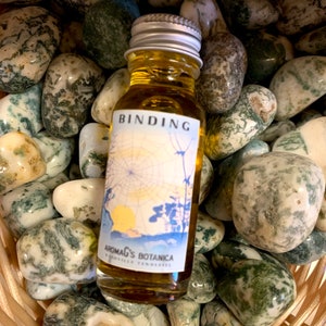 May include: A small glass bottle with a silver lid and a label that says "BINDING" and "Aromatherapy's Botanica". The bottle is filled with a yellow liquid and is sitting on a bed of green and white stones.