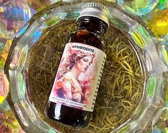 Könnte beinhalten: Eine kleine braune Glasflasche mit einem silbernen Deckel, die eine Flüssigkeit enthält. Die Flasche hat ein Etikett mit einer Zeichnung einer Frau und dem Wort "APHRODITE" darauf gedruckt. Das Etikett enthält auch den Text "SPELLCASTING BY AROMAGS.COM".