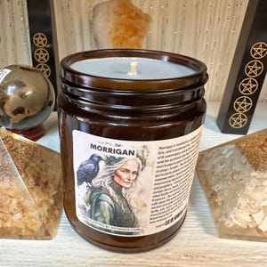 May include: A brown glass jar candle with a label featuring the name "Morrigan" and an illustration of a woman with a raven. The candle has a light gray wax and a wick. The label also includes text about the candle's purpose.