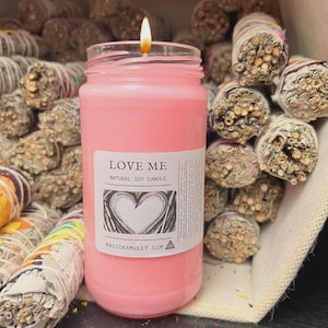 May include: A lit pink natural soy candle in a glass jar with a label that reads "LOVE ME" and a heart design. The candle is surrounded by bundles of dried sage. The website address is printed at the bottom of the label.