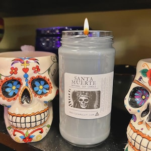 May include: A lit, gray candle in a glass jar with a label that reads "SANTA MUERTE NATURAL SOY CANDLE." The candle is next to a decorative skull-shaped ceramic container with colorful floral designs. The candle's flame is bright.