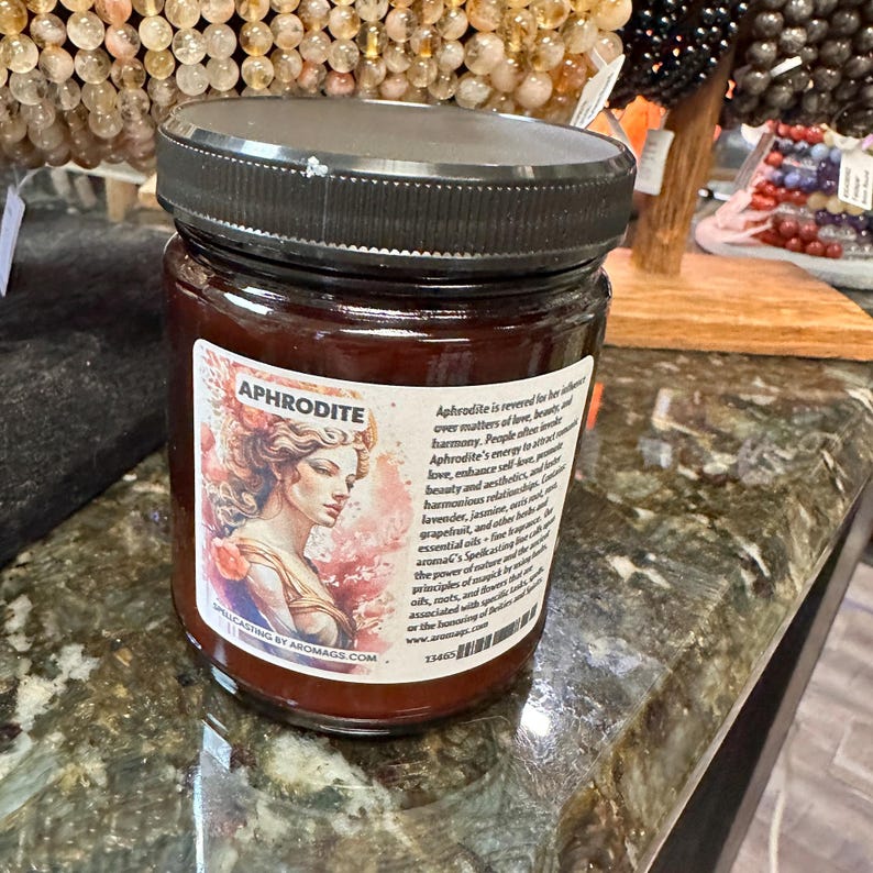 May include: A brown glass jar with a black lid and a label that says "APHRODITE" and has a picture of a woman. The label also includes text about the goddess Aphrodite and her association with love, beauty, and harmony. The jar is filled with a red liquid.