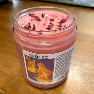 May include: Pink candle in a glass jar with a label that says "SEDUCE". The label also includes text about bringing out seduction and the ingredients used in the candle. The candle has dried rose petals on top.