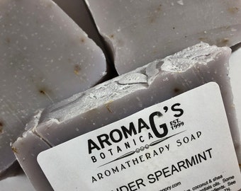 Könnte beinhalten: Drei Stück Lavendel-Minz-Aromatherapie-Seife von AromaG's Botanica, gegründet 1999. Die Seife ist helllila und hat eine gesprenkelte Optik. Die Seife ist in einer weißen Schachtel mit dem Firmenlogo und Produktinformationen verpackt.