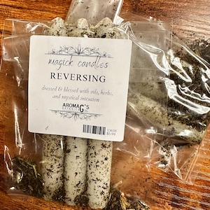 May include: Three white candles with green herbs and a label that reads "magick candles REVERSING dressed & blessed with oils, herbs, and mystical intention AROMAC'S BOTANICA 13638 $5.99"