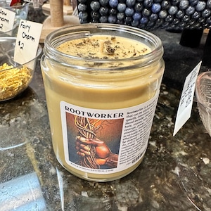 May include: A glass jar candle with a brown label that reads "Rootworker" and includes a description of the candle's purpose. The candle is topped with dried herbs and has a brown wax base.
