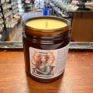 May include: A brown glass candle jar with a yellow wax candle inside. The jar has a label with a drawing of a woman with red hair and the word "Brigid" on it.