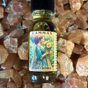 May include: A small glass bottle with a black cap, filled with a golden liquid. The label reads "LAMMAS" and features a woman holding a sunflower. The text "AROMAG'S BOTANICA NASHVILLE TENNESSEE" is also visible. The bottle rests on a bed of rough, light-brown stones.