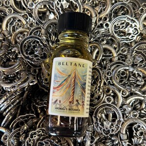 May include: A small glass bottle with a black cap, filled with a golden liquid. The label reads "Beltane" and features a colorful illustration of a maypole. The bottle sits on a bed of silver charms.