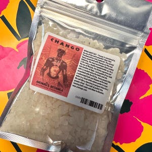 May include: A clear plastic bag filled with bath salts. The label reads "CHANGO" with an illustration of a figure. Text on the label describes the ritual use of the bath salts. The brand name "Aromag's Botanica" is also visible.