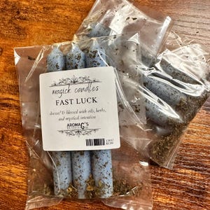 May include: Three blue candles with herbs and oils, packaged in a clear plastic bag. The label reads "magick candles FAST LUCK" and "dressed & blessed with oils, herbs, and mystical intention" with the company name "AROMAG'S BOTANICALS" and a price tag of $5.99.
