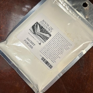 May include: A sealed, clear plastic bag containing white sachet powder. The bag has a label with the text "BOOK OF SHADOWS" and "SACHET POWDER". The label also includes text about the product's use with herbs, oils, and flowers.