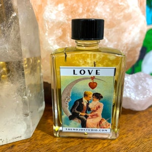May include: A small glass bottle with a black cap filled with a yellow liquid. The bottle has a label that says "LOVE" and features a vintage illustration of a couple sitting on a crescent moon with a red heart hanging from a string.