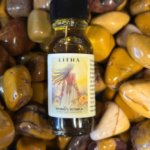 May include: A clear glass bottle with a black cap, filled with a golden liquid. The label reads "LITHA" and features an illustration of a woman. The bottle is surrounded by yellow and brown stones, with the text "AROMAG'S BOTANICA" at the bottom.